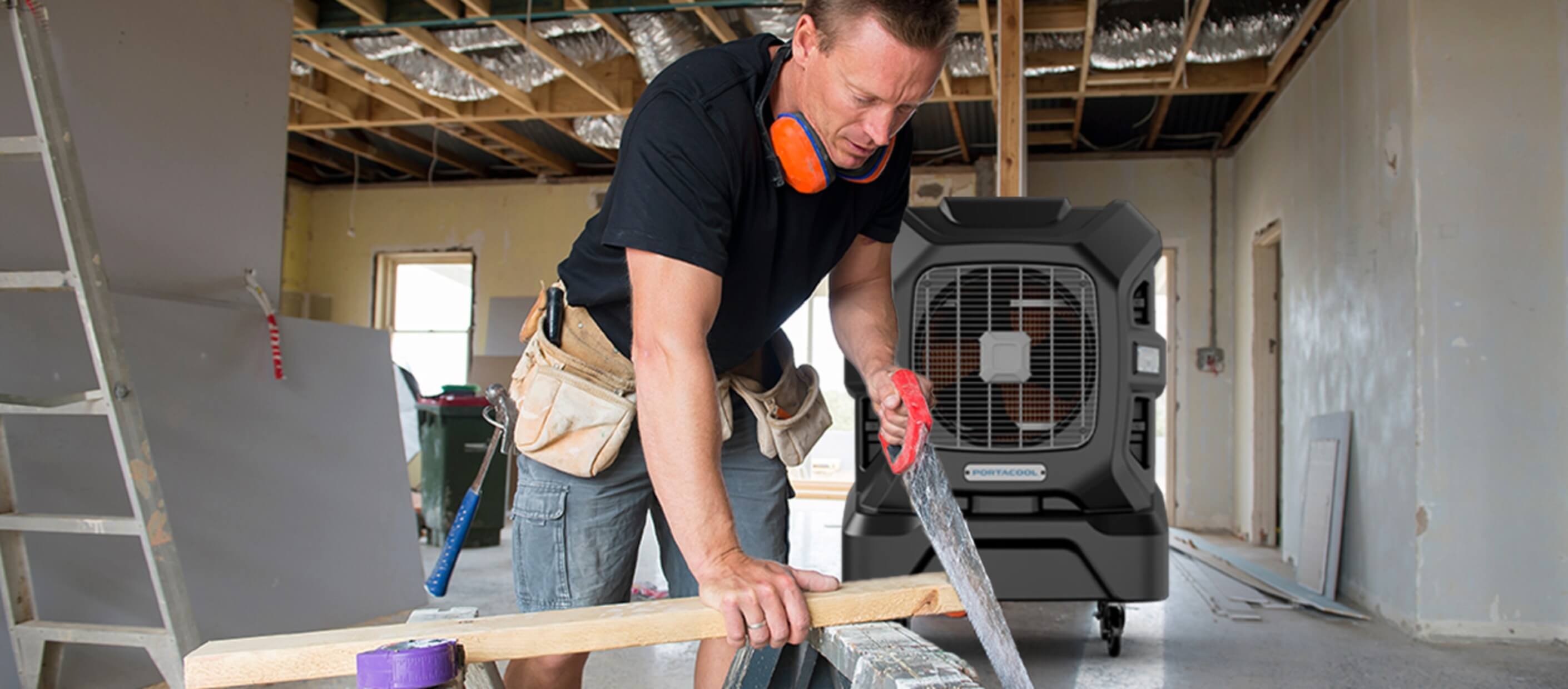 Construction worker cutting wood on sawhorse, wearing a tool belt and earmuffs. Exposed ceiling beams and drywall indicate an active construction site.