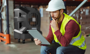 A worker in a vest and hard hat sits in a warehouse. He's wondering what the difference between air conditioning and evaporative cooling is.