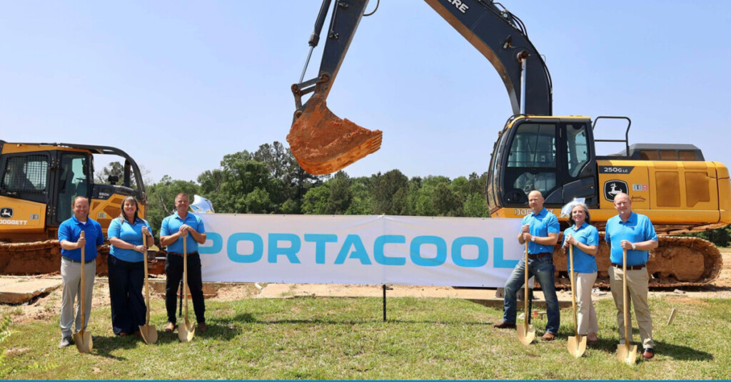 Portacool sign about shoulder height on a grass field between groups of Portacool leadership members, in front of industrial machinery breaking ground for the new facility.