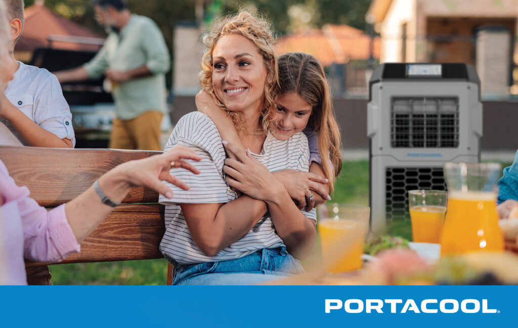 People relax in their backyard, sitting at a table with food and drink, kept comfortable by a Portacool evaporative cooler in the background.