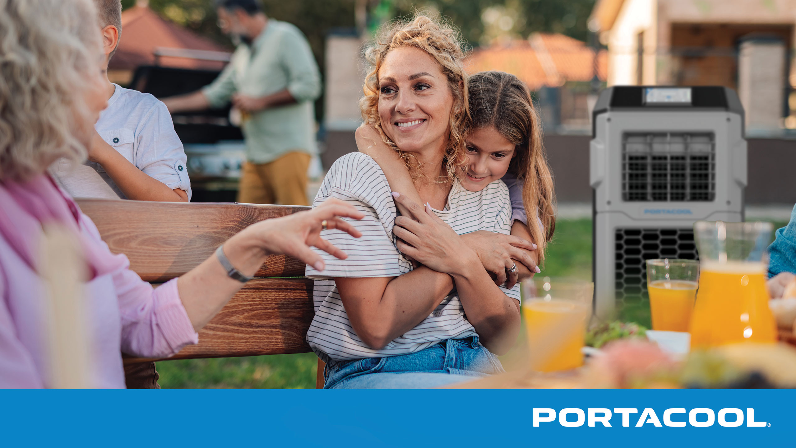 People relax in their backyard, sitting at a table with food and drink, kept comfortable by a Portacool evaporative cooler in the background.