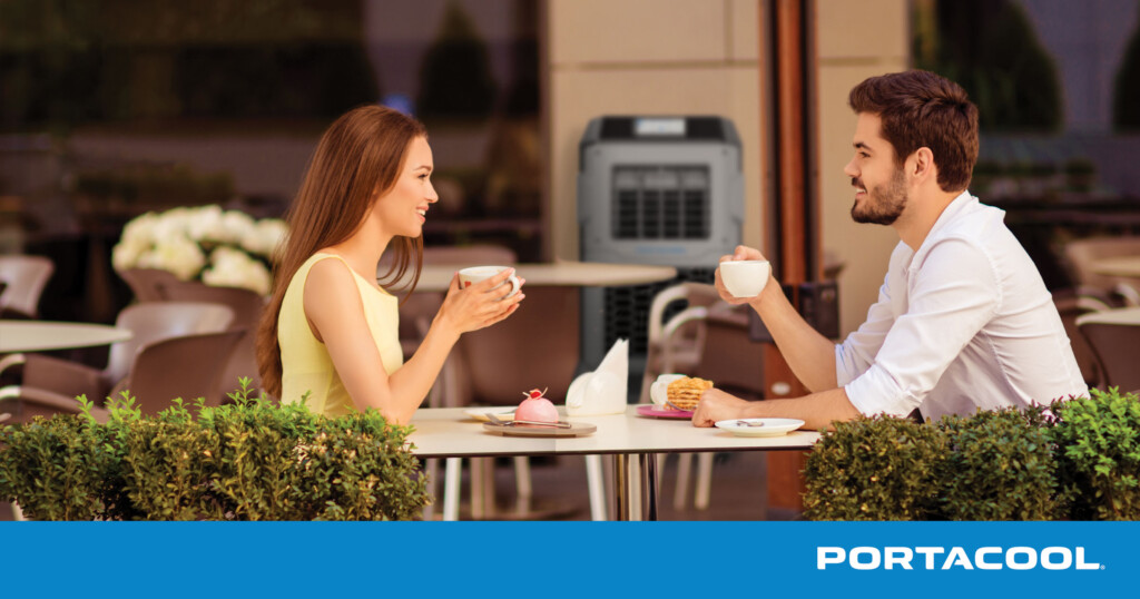 a man and woman sitting at a table with coffee cups, with a Portacool evaporative cooler in the background