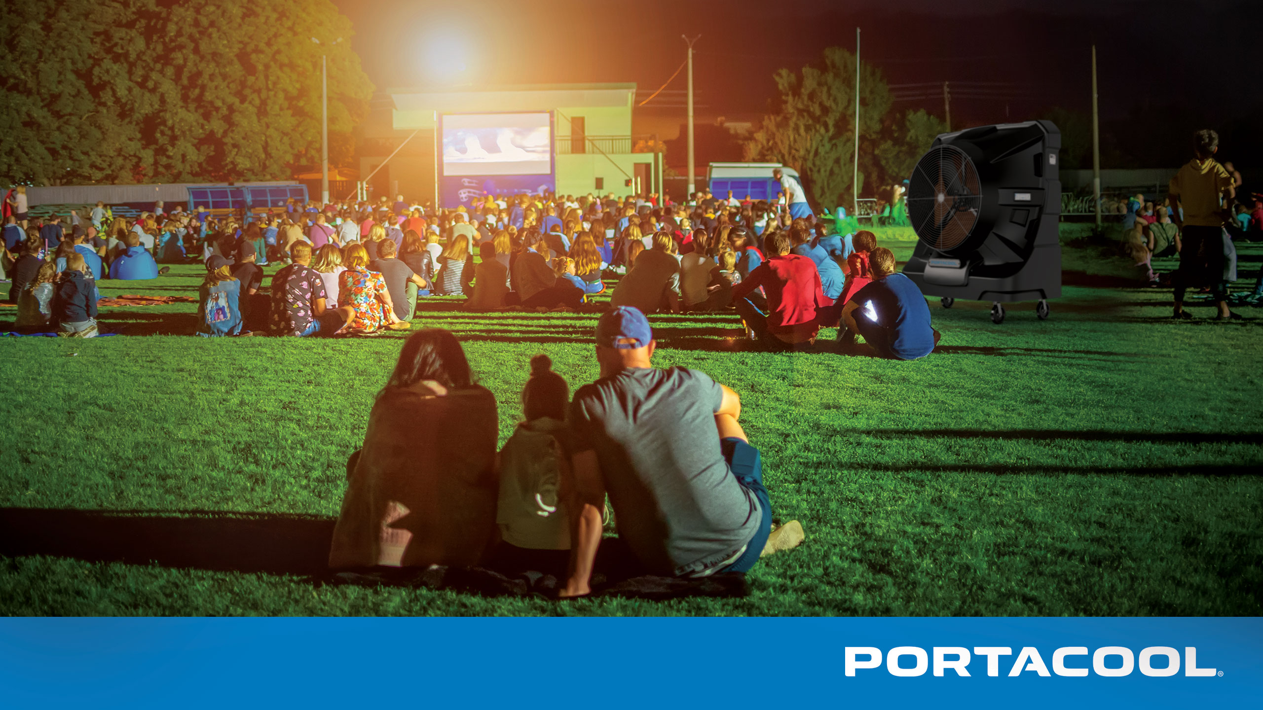 A family gathers to watch a movie outdoors on a hot summer evening during a community event, kept cool by a Portacool portable evaporative cooler.